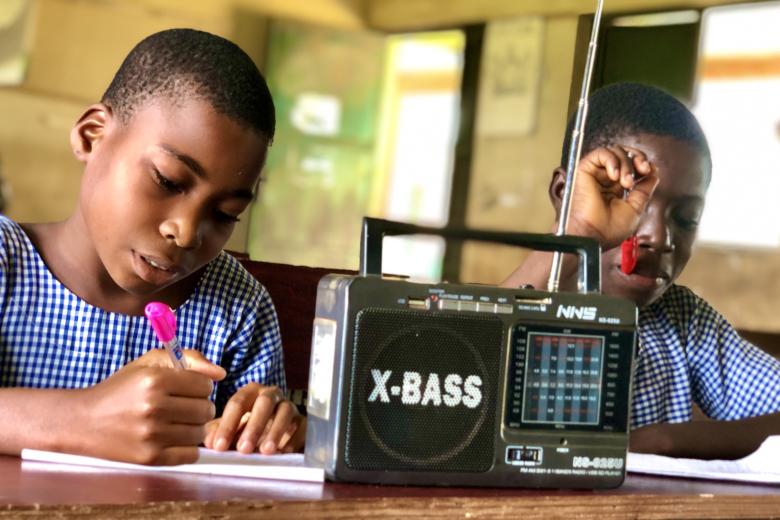 Two African girls in gingham school uniforms write on paper at a desk with a radio in front of them that says X-Bass