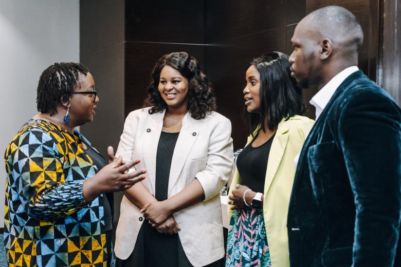 A Black woman in a colorful shirt speaks as two Black women and one Black man listen smiling. All are standing close together in conversation.