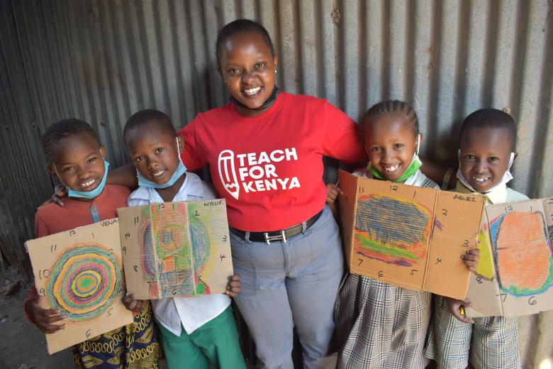 A young Black woman in a red Teach For Kenya t-shirt stands with her arms around the shoulders o four young Black children who are holding drawings. All are smiling