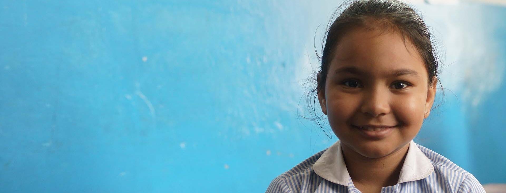 Close up of a young South Asian girl smiling with a painted blue wall in the background