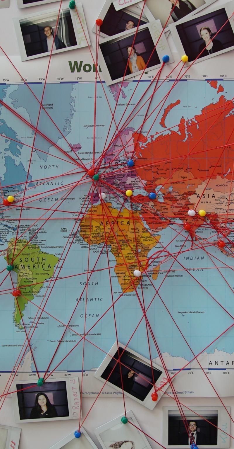 A map of the world on a wall with photographs surrounding it and red threads running from the photos to localities all over the map