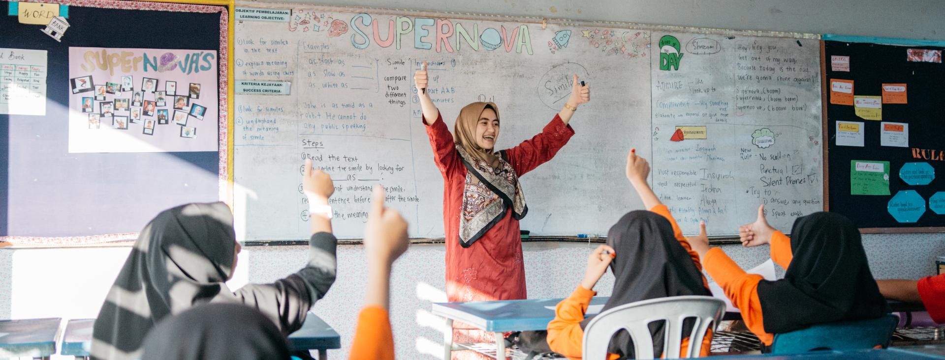A female teacher in a red dress and head scarf stands in front of a white board facing her students in desks. Everyone raises their arms giving a thumbs-up