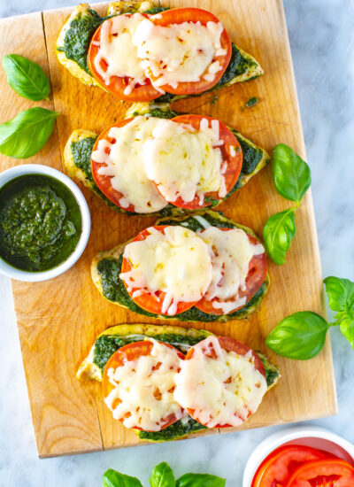 Four chicken cutlets on a butcher block topped with pesto, tomato and mozzarella cheese.