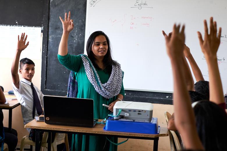 A female teacher in India and her students raise their hands