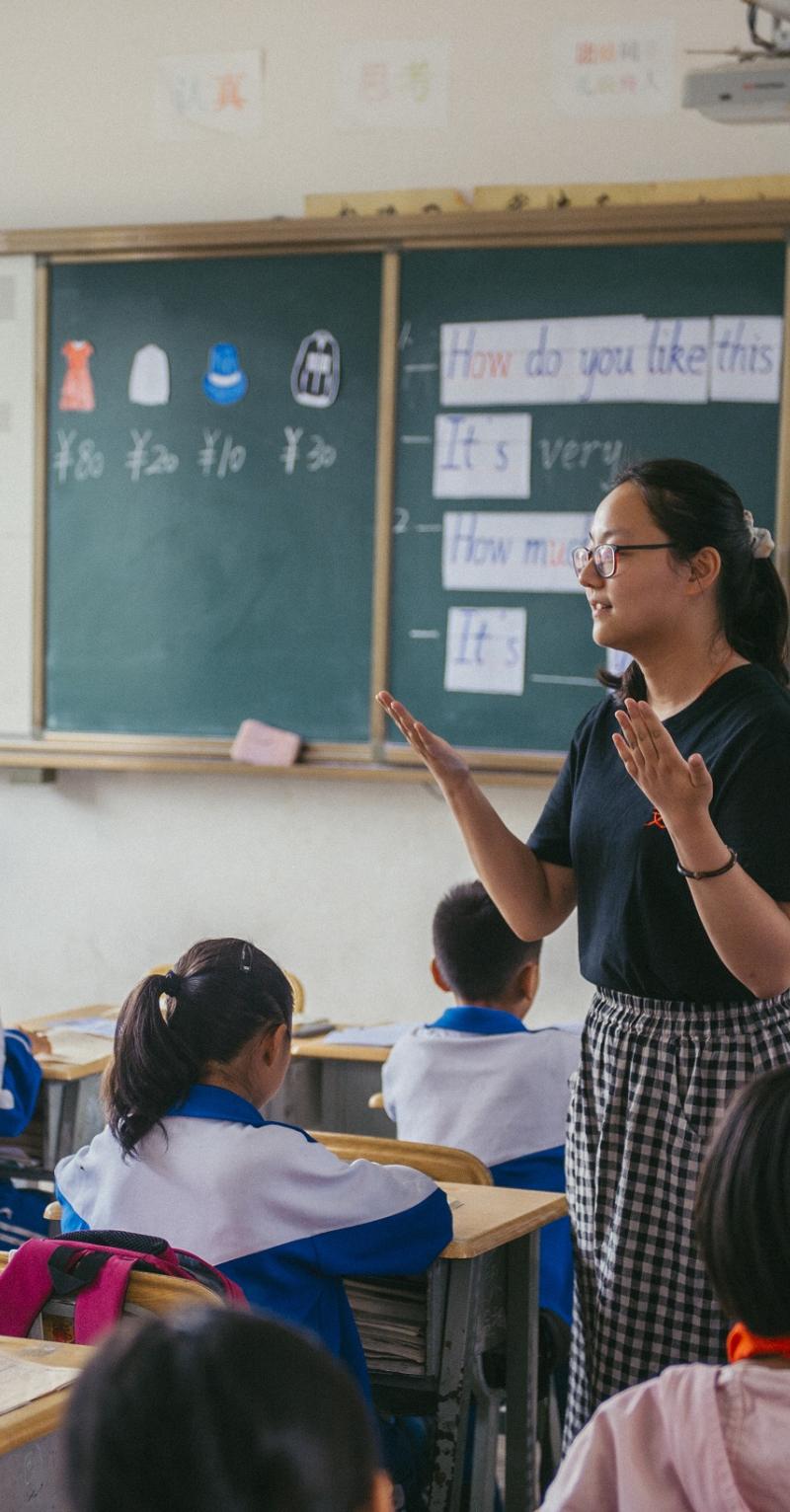 A young Asian woman stands in a classroom in with a chalkboard behind her among students wearing white shirts seated in desks 
