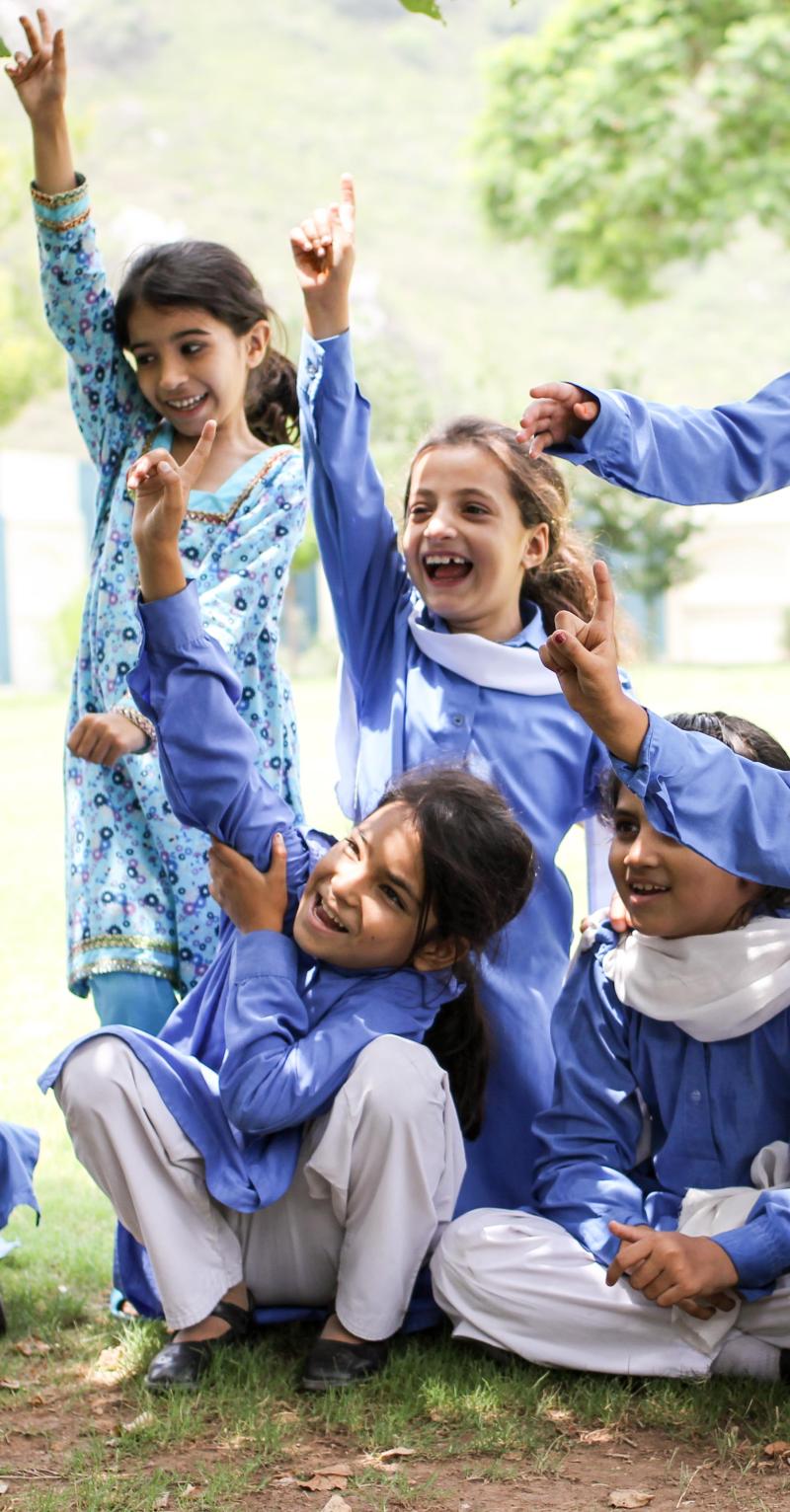 A group of girls in blue tunics with white sashes smile broadly with their hands in the air. Some are seated and some are standing.