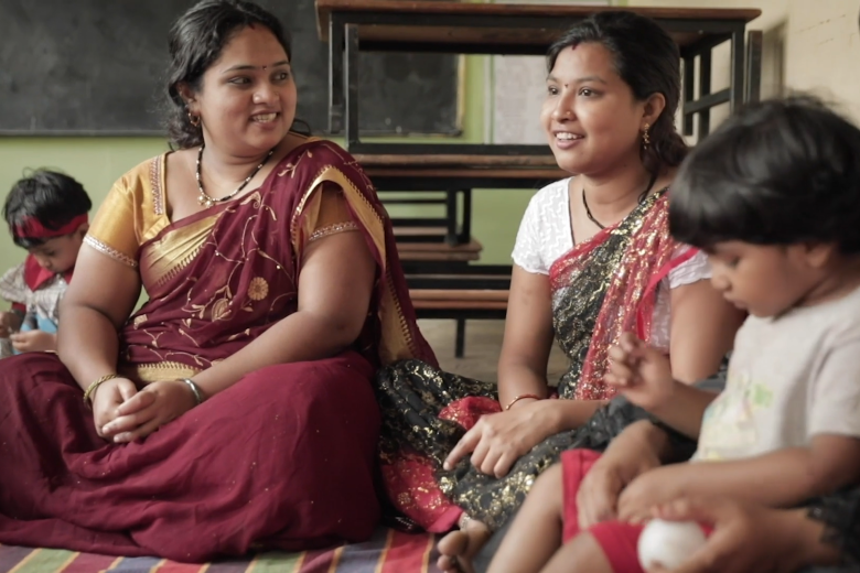 Two women in saris smile while seated on the floor next to a small child