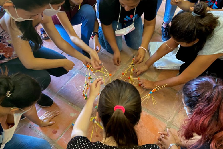 A group of adults crouch in a circle on the floor while constructing something together out of pick-up sticks